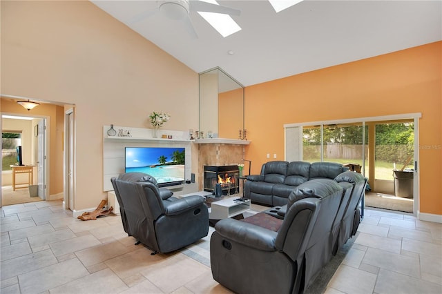 living room with ceiling fan, a skylight, a tiled fireplace, and high vaulted ceiling