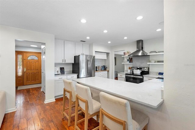 kitchen with dark hardwood / wood-style floors, white cabinets, kitchen peninsula, wall chimney range hood, and appliances with stainless steel finishes