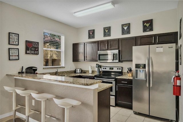 kitchen featuring light tile patterned flooring, kitchen peninsula, light stone countertops, stainless steel appliances, and a kitchen bar