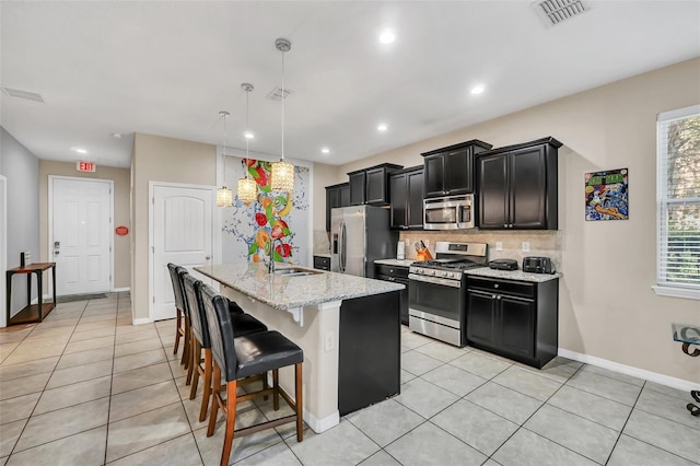 kitchen featuring pendant lighting, light stone counters, an island with sink, backsplash, and appliances with stainless steel finishes