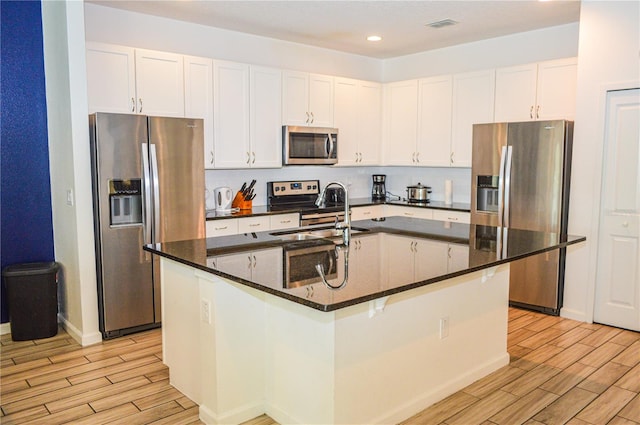 kitchen with white cabinetry, stainless steel appliances, dark stone counters, and a kitchen island with sink