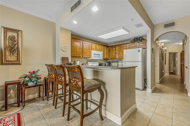 kitchen with light tile patterned floors, ornamental molding, kitchen peninsula, white appliances, and a kitchen breakfast bar