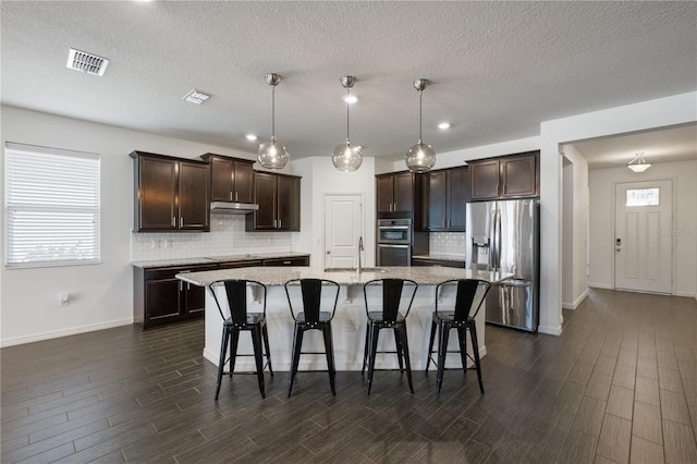 kitchen with a kitchen breakfast bar, stainless steel appliances, dark hardwood / wood-style floors, hanging light fixtures, and an island with sink