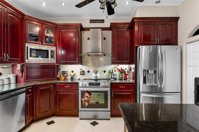 kitchen featuring ceiling fan, wall chimney range hood, appliances with stainless steel finishes, dark stone countertops, and crown molding