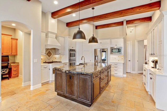 kitchen with appliances with stainless steel finishes, dark stone countertops, pendant lighting, beam ceiling, and a center island with sink