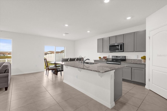 kitchen with light stone counters, a kitchen island with sink, gray cabinets, stainless steel appliances, and a kitchen bar