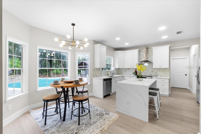 kitchen featuring wall chimney range hood, light hardwood / wood-style flooring, stainless steel dishwasher, a center island, and white cabinetry