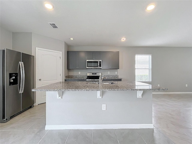 kitchen featuring appliances with stainless steel finishes, a kitchen island with sink, light stone countertops, and gray cabinets