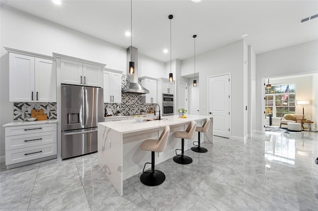 kitchen featuring an island with sink, wall chimney exhaust hood, hanging light fixtures, a breakfast bar area, and appliances with stainless steel finishes