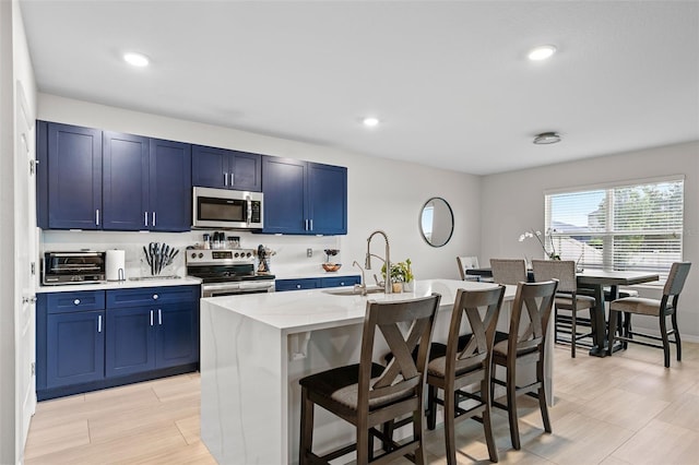 kitchen with light stone counters, a kitchen island with sink, a kitchen breakfast bar, stainless steel appliances, and blue cabinetry