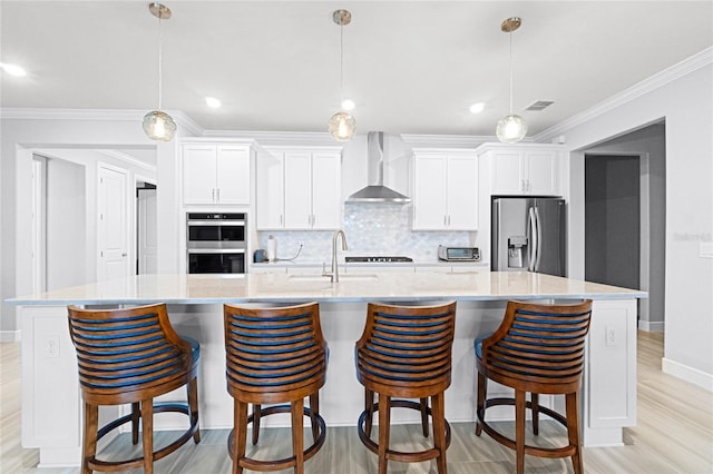 kitchen with a large island with sink, appliances with stainless steel finishes, white cabinetry, and wall chimney range hood