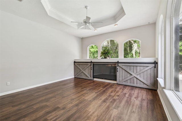 empty room featuring a barn door, a raised ceiling, dark hardwood / wood-style flooring, ceiling fan, and ornamental molding