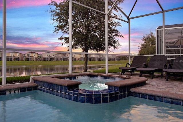 pool at dusk with an in ground hot tub, a water view, glass enclosure, and a patio area