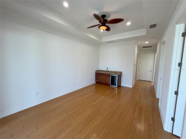 empty room with ceiling fan, a raised ceiling, ornamental molding, beverage cooler, and light hardwood / wood-style flooring