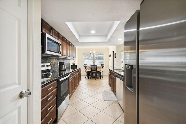 kitchen with a tray ceiling, an inviting chandelier, backsplash, appliances with stainless steel finishes, and light tile patterned floors