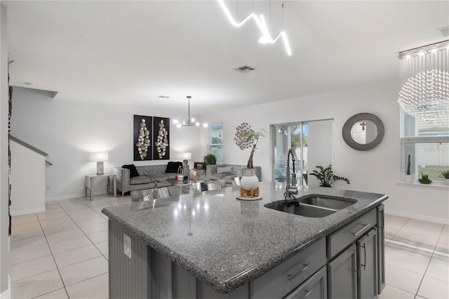 kitchen featuring sink, dark stone counters, light tile patterned floors, a kitchen island with sink, and a notable chandelier