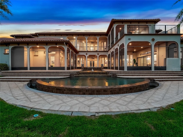 back house at dusk featuring a pool with hot tub, a balcony, and a patio