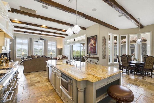 kitchen with beamed ceiling, an island with sink, sink, stainless steel appliances, and light stone countertops