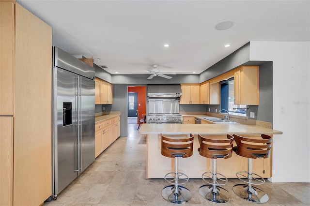 kitchen with wall chimney range hood, high quality appliances, kitchen peninsula, ceiling fan, and a breakfast bar area