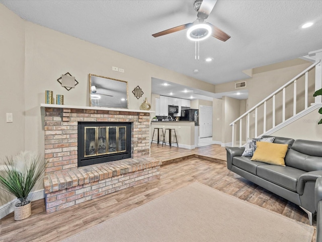 living room featuring ceiling fan, light hardwood / wood-style floors, a textured ceiling, and a brick fireplace