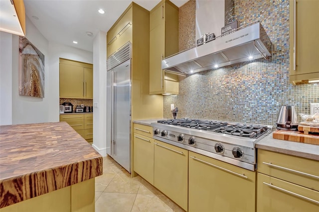 kitchen with wall chimney range hood, light tile patterned floors, appliances with stainless steel finishes, backsplash, and butcher block counters