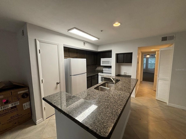 kitchen with a center island with sink, white appliances, light tile patterned floors, dark stone counters, and sink