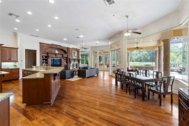 kitchen with light stone counters, ornamental molding, a center island with sink, dark wood-type flooring, and ornate columns