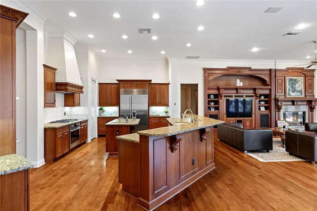 kitchen with custom range hood, an island with sink, light stone countertops, stainless steel appliances, and hardwood / wood-style flooring