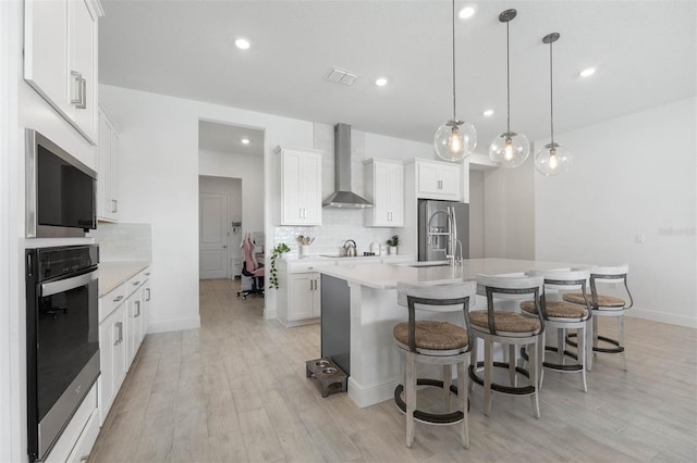 kitchen featuring light wood-type flooring, wall chimney range hood, pendant lighting, white cabinets, and an island with sink