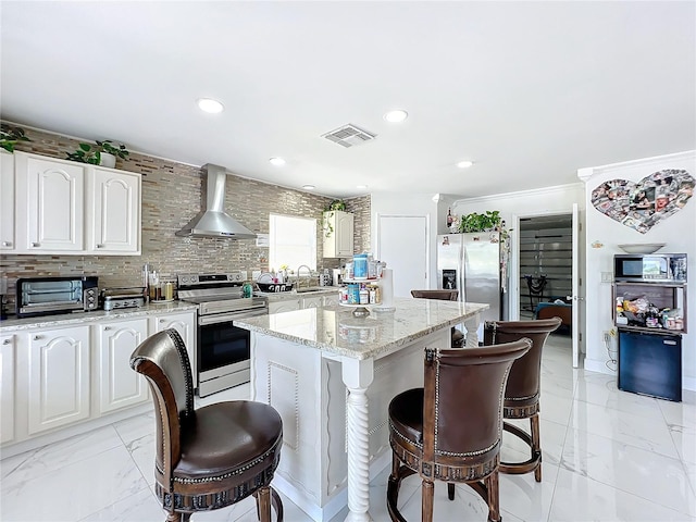 kitchen with light stone counters, stainless steel appliances, wall chimney range hood, a kitchen island, and white cabinetry