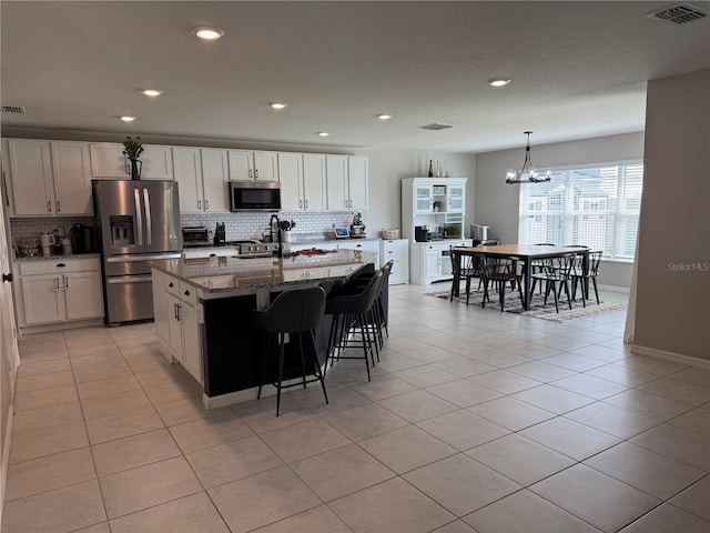 kitchen featuring appliances with stainless steel finishes, white cabinets, light tile patterned flooring, stone counters, and a center island with sink