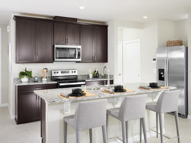 kitchen with appliances with stainless steel finishes, light tile patterned floors, an island with sink, and light stone counters