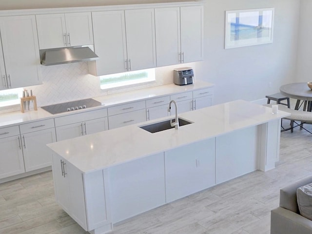 kitchen featuring black electric stovetop, backsplash, a kitchen island with sink, sink, and white cabinetry