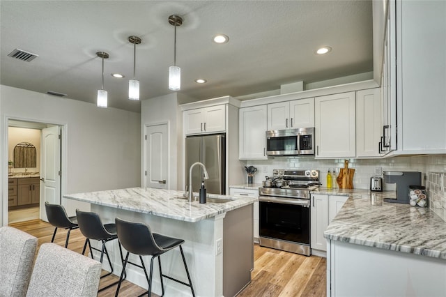 kitchen featuring appliances with stainless steel finishes, light hardwood / wood-style floors, white cabinets, decorative light fixtures, and a kitchen island with sink