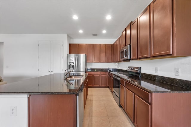 kitchen featuring appliances with stainless steel finishes, sink, dark stone counters, light tile patterned floors, and a kitchen island with sink