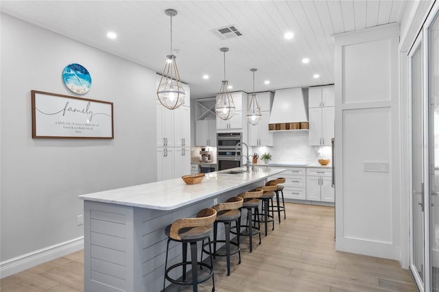 kitchen with custom range hood, light stone counters, stainless steel double oven, decorative light fixtures, and a large island