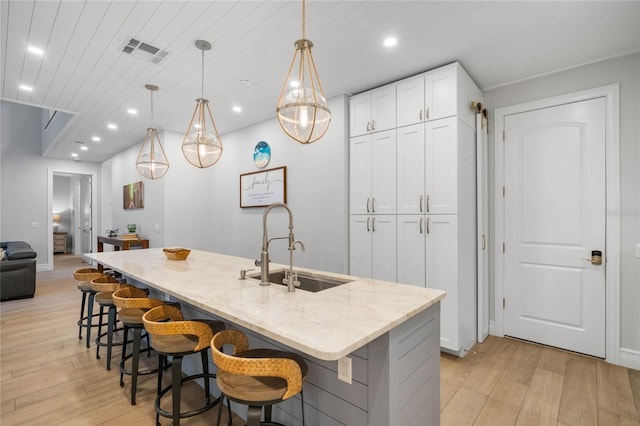 kitchen with hanging light fixtures, light stone counters, white cabinets, light wood-type flooring, and a center island with sink