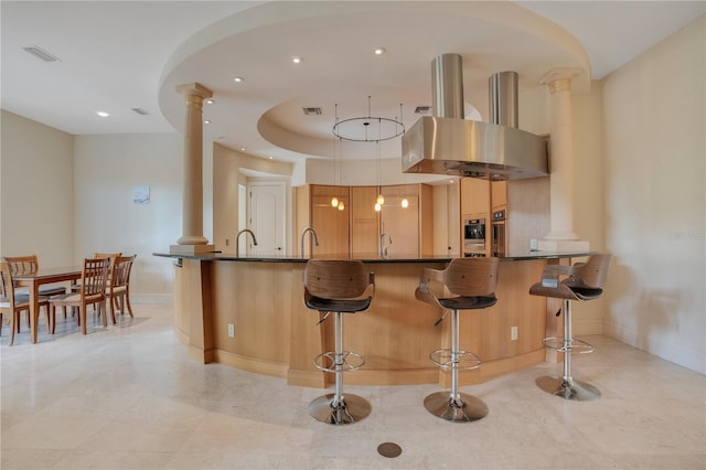 kitchen with a raised ceiling, a breakfast bar area, kitchen peninsula, island exhaust hood, and decorative columns