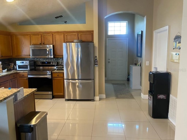 kitchen featuring light stone countertops, light tile patterned floors, appliances with stainless steel finishes, high vaulted ceiling, and tasteful backsplash