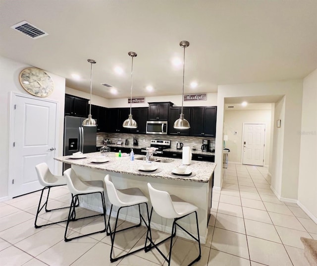 kitchen with stainless steel appliances, pendant lighting, light stone counters, decorative backsplash, and a kitchen island with sink