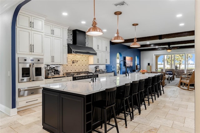 kitchen with a large island, beam ceiling, white cabinetry, hanging light fixtures, and wall chimney exhaust hood