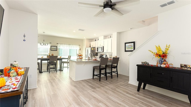 kitchen with white cabinets, light wood-type flooring, stainless steel appliances, a kitchen breakfast bar, and ceiling fan with notable chandelier