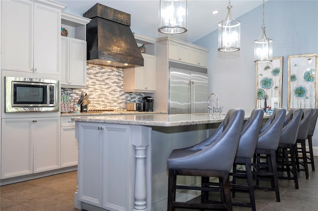 kitchen featuring premium range hood, a center island with sink, white cabinetry, and built in appliances