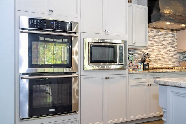 kitchen with white cabinets, wall chimney exhaust hood, backsplash, appliances with stainless steel finishes, and light stone countertops