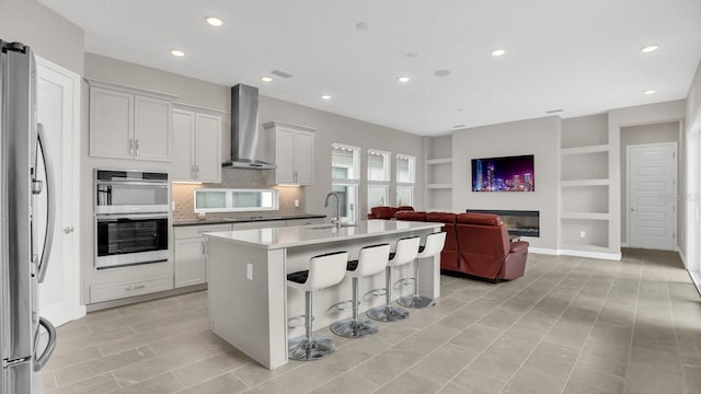 kitchen featuring wall chimney exhaust hood, stainless steel appliances, white cabinets, and a kitchen island with sink