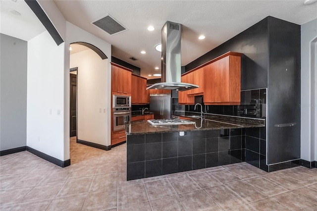 kitchen with island exhaust hood, visible vents, dark stone countertops, built in appliances, and a peninsula