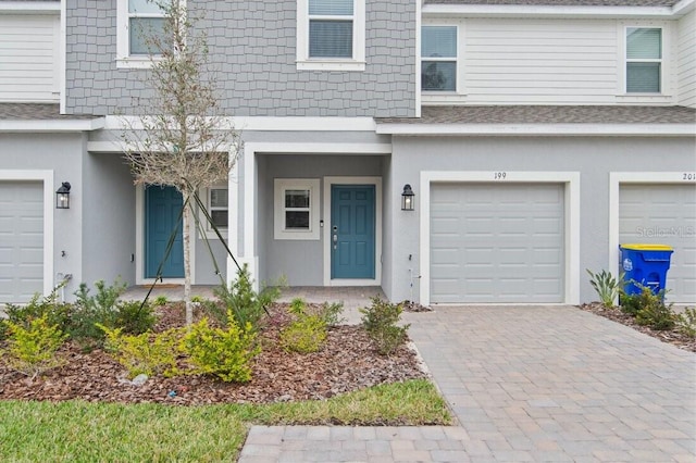 property entrance with stucco siding, decorative driveway, an attached garage, and a shingled roof