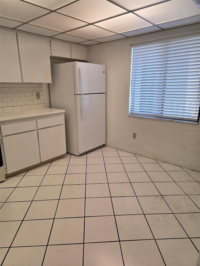 kitchen with a paneled ceiling, backsplash, white refrigerator, light tile patterned floors, and white cabinetry