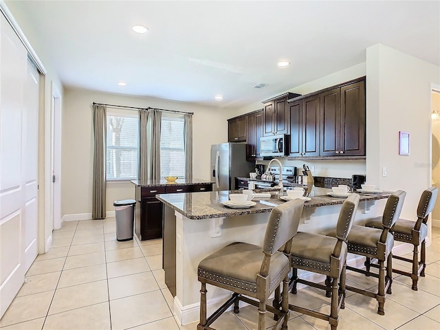 kitchen featuring dark brown cabinetry, light tile patterned flooring, stainless steel appliances, a breakfast bar, and dark stone counters