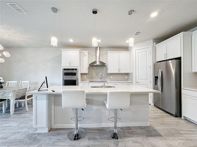 kitchen with appliances with stainless steel finishes, wall chimney range hood, an island with sink, and decorative light fixtures